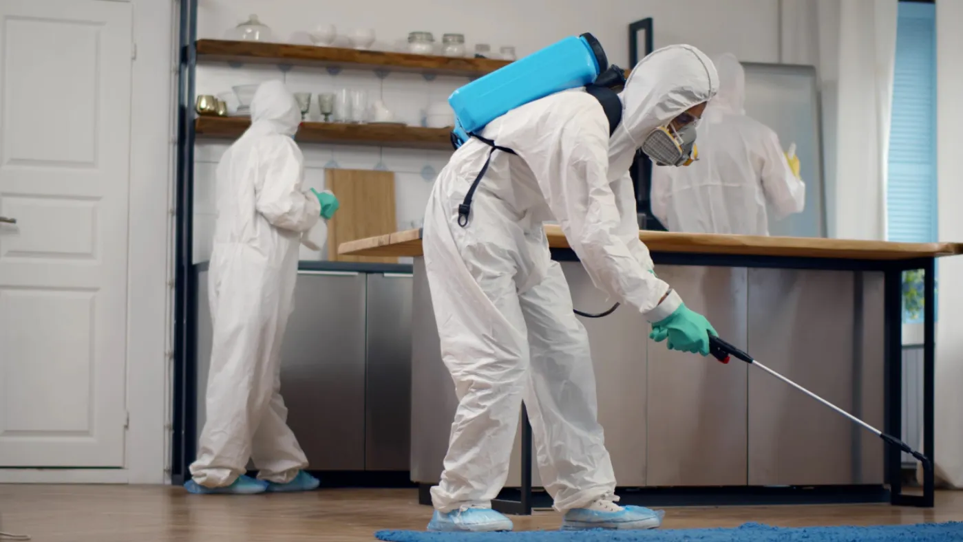 A team of pest control professionals in full hazmat suits and respirator masks disinfects a residential property, with one exterminator using a backpack pressure sprayer on the floor.