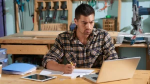Handyman managing business tasks on a laptop in a workshop surrounded by tools and equipment.
