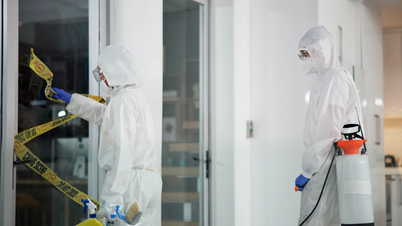 Crime scene cleaners in full protective suits disinfecting an indoor area with caution tape and sanitation equipment.