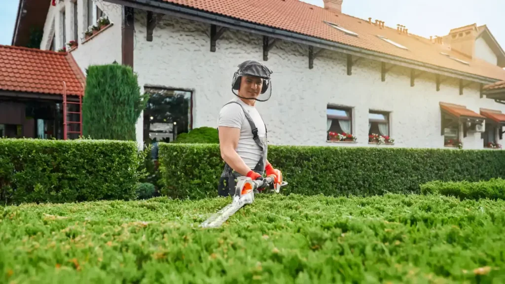 landscaping pro in uniform, glasses, and gloves pruning bushes in garden.