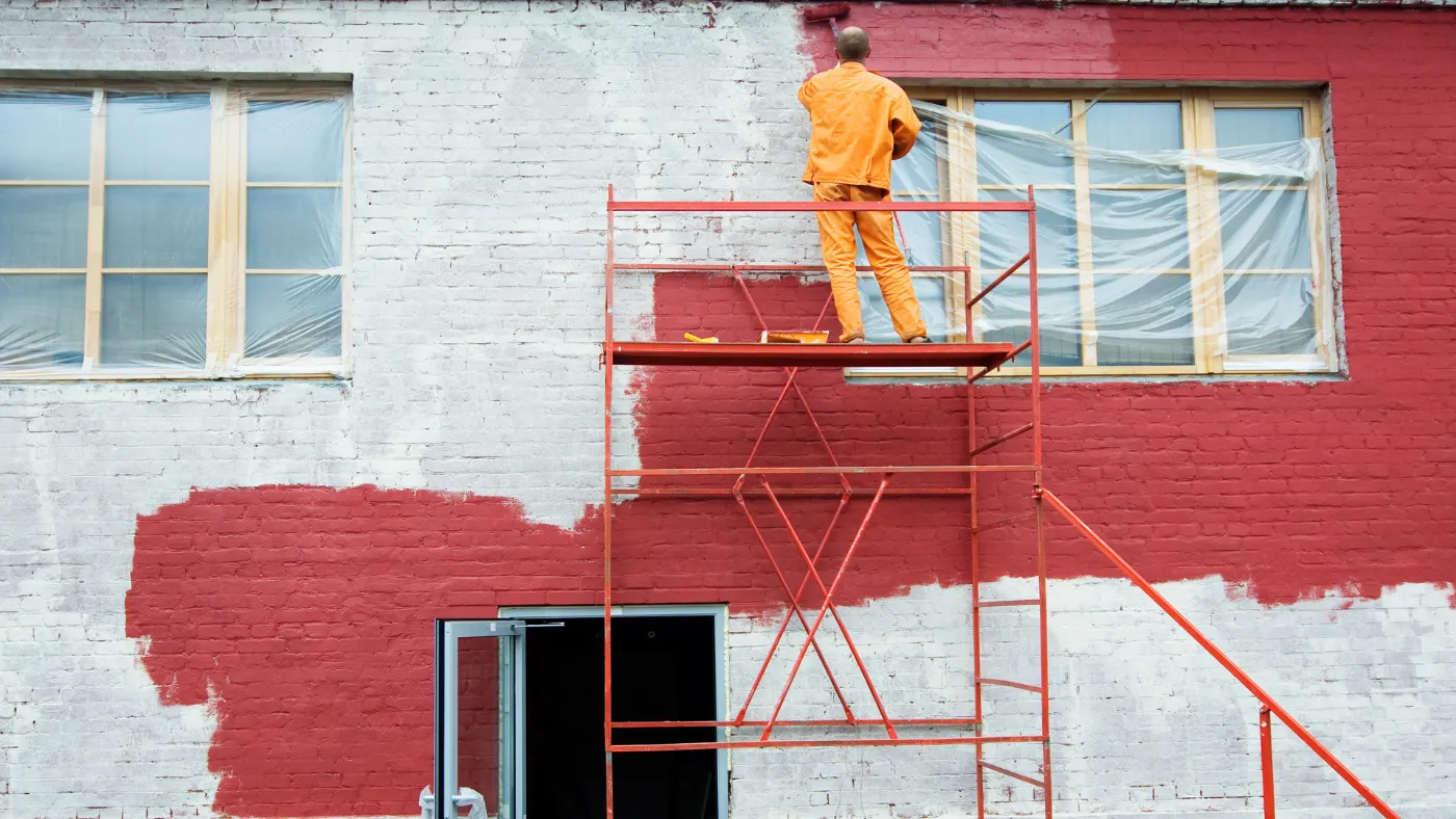 Painter on scaffolding applying red paint to a brick building with windows covered in plastic