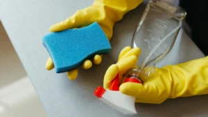 Hands in yellow gloves holding a blue sponge and spray bottle.