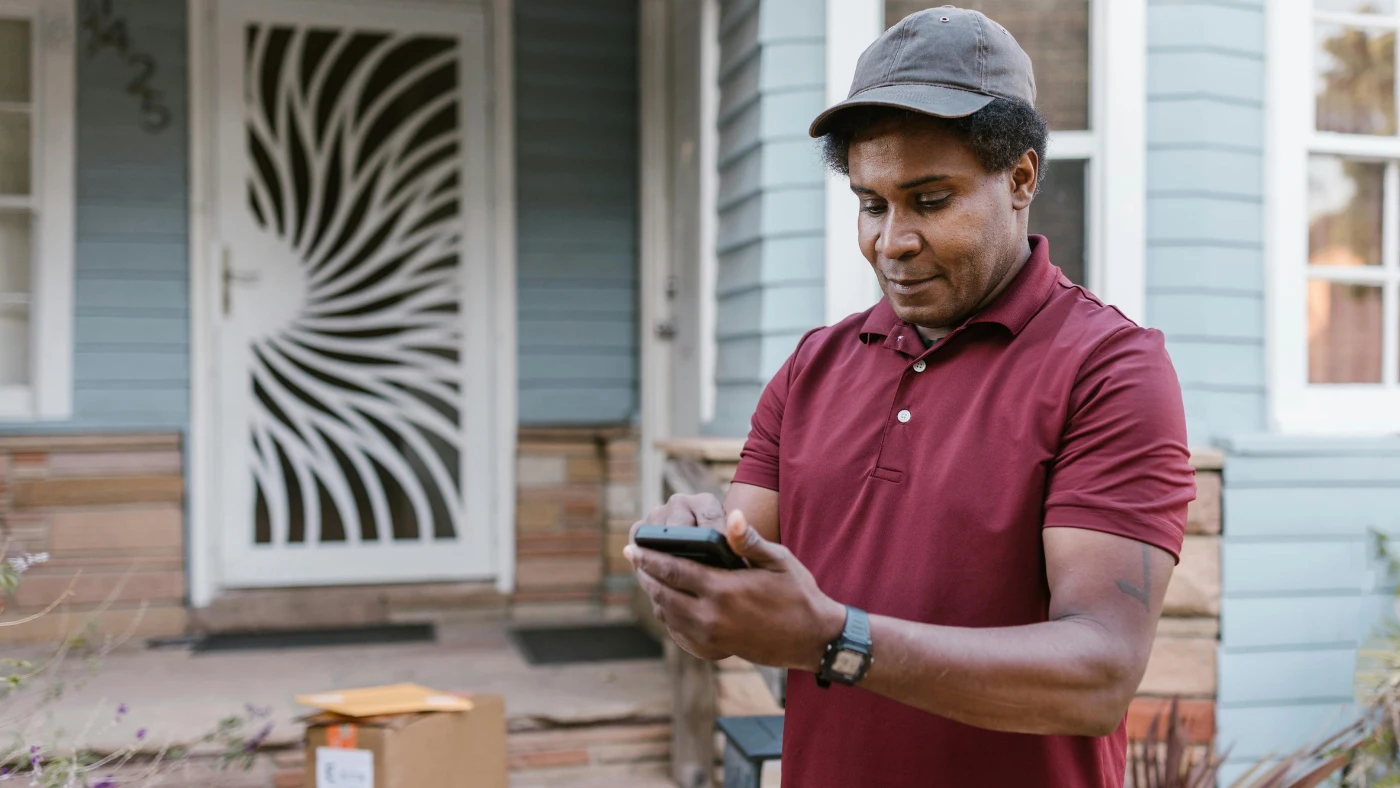 Delivery worker checking a smartphone outside a house with packages nearby.