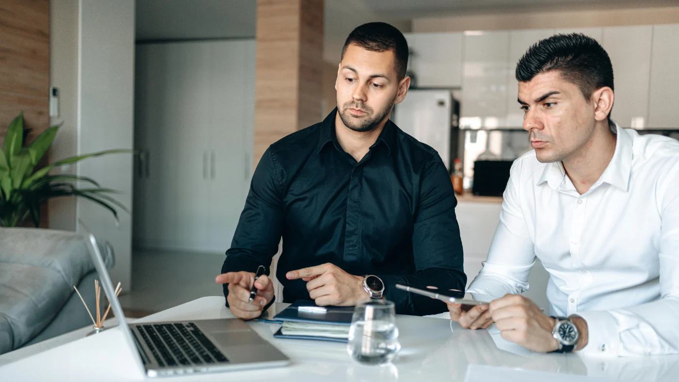 two men sitting together, both looking at a laptop screen and discussing work