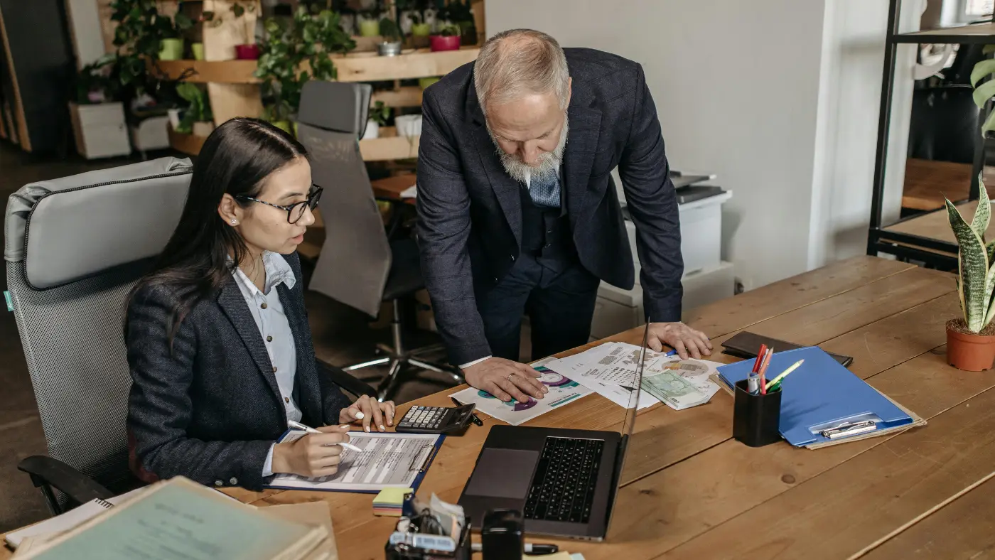 Man standing beside a woman sitting at a desk, both discussing while looking at a laptop screen