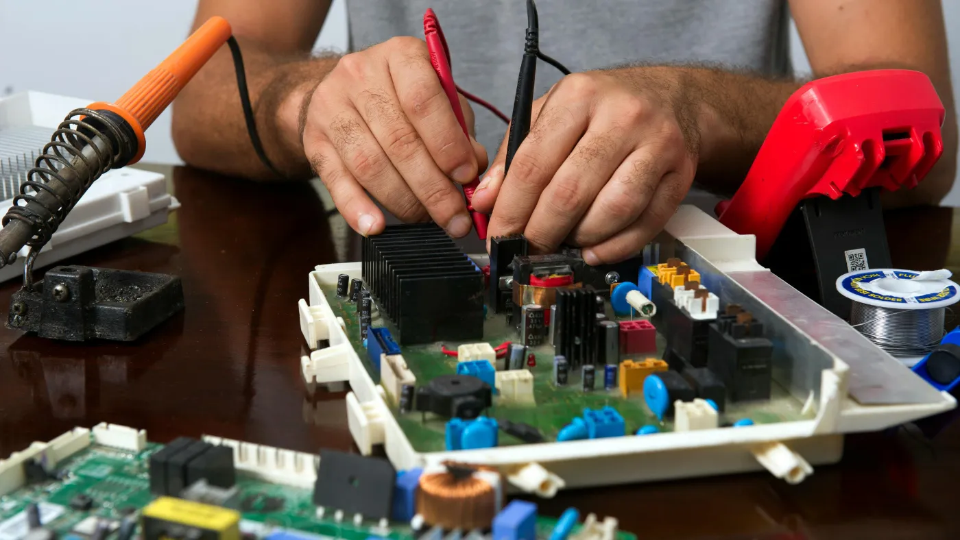 Technician testing an appliance control board with a multimeter and soldering tools