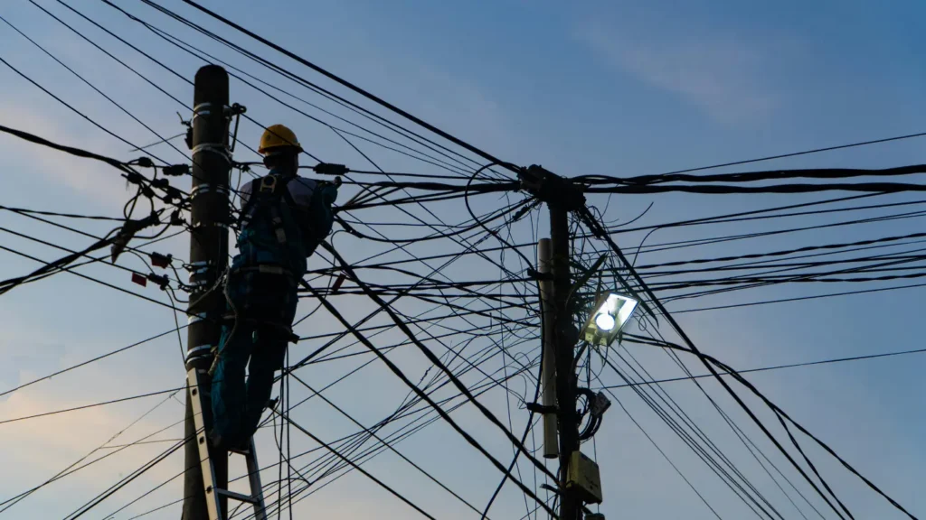 Worker on a ladder repairing dense overhead power lines on utility poles.
