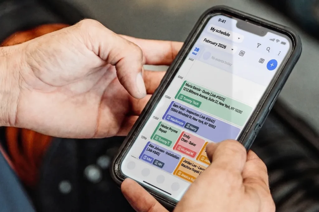 Two hands hold a smartphone displaying the Housecall Pro mobile schedule. The calendar view shows color-coded job blocks for multiple technicians.