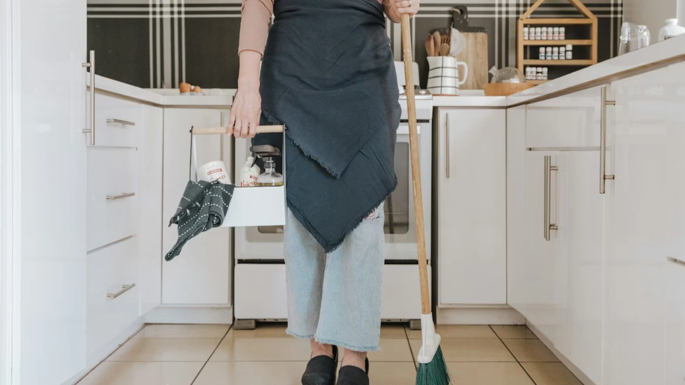 Professional house cleaner holding a broom and cleaning supplies in a modern kitchen, representing residential and commercial cleaning service pricing
