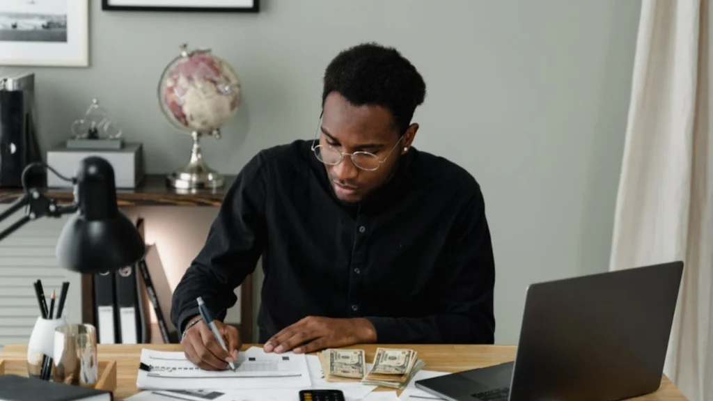 Small business owner calculating payroll at desk with laptop, cash, and paperwork, representing DIY payroll management for small businesses
