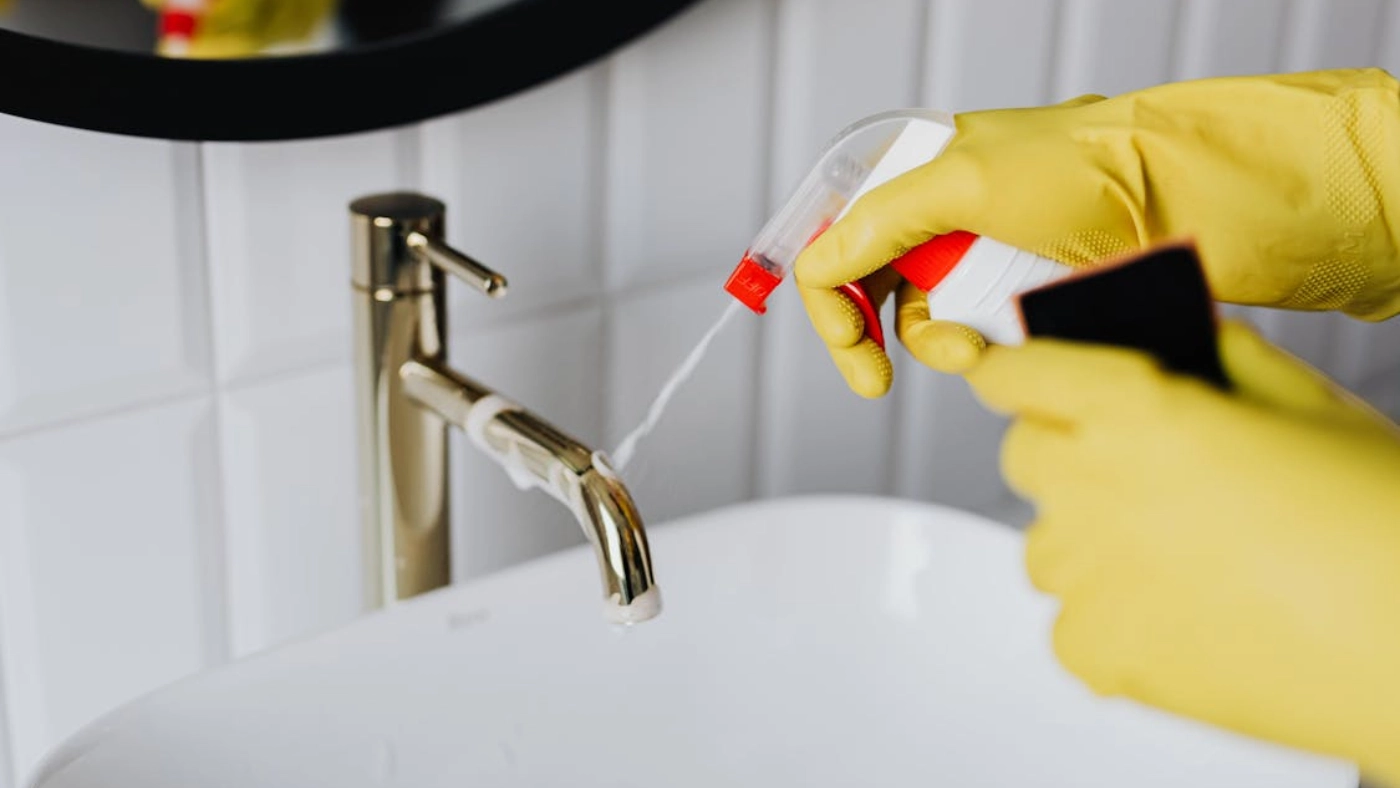 Person cleaning bathroom sink faucet with spray cleaner and gloves