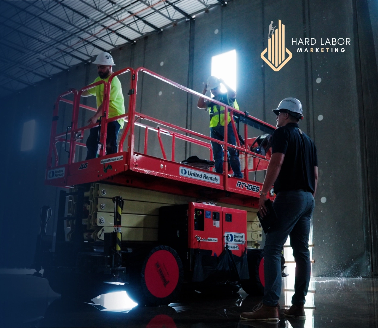 Three construction workers on a red scissor lift in a warehouse, wearing hard hats and high-visibility vests; Hard Labor Marketing logo in the corner.