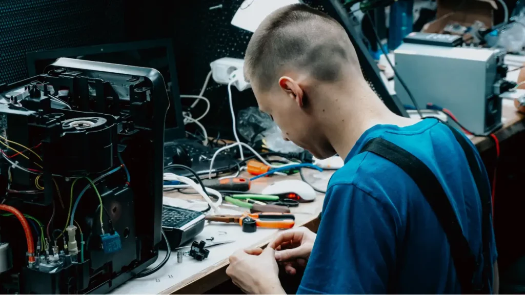 Repair technician working on the internal components of a disassembled small appliance at a cluttered workbench, with tools spread nearby.