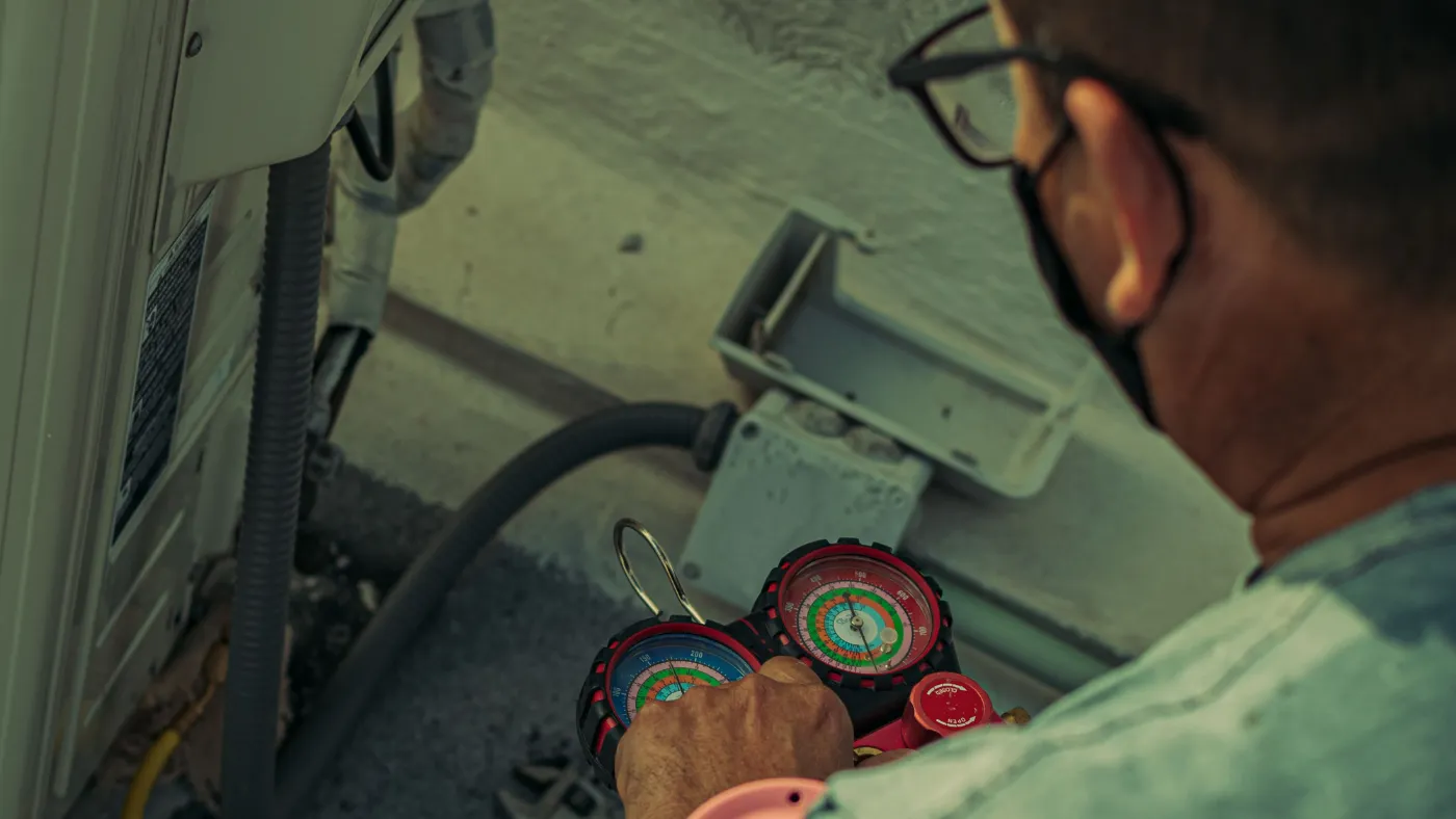 HVAC technician using red and blue refrigerant manifold gauges to service an outdoor air conditioning condenser unit.