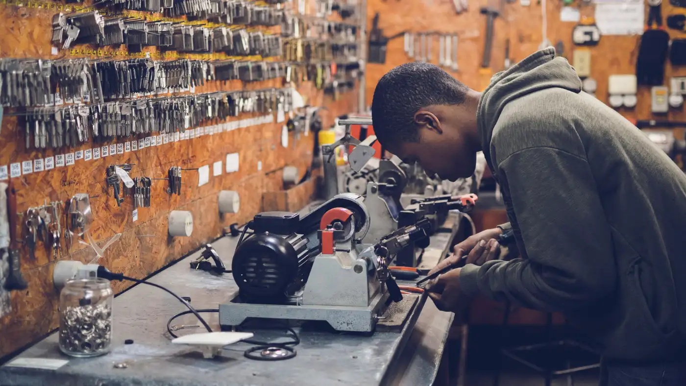 Person in a gray hoodie works at a bench grinder on a cluttered workshop workbench filled with tools and parts.
