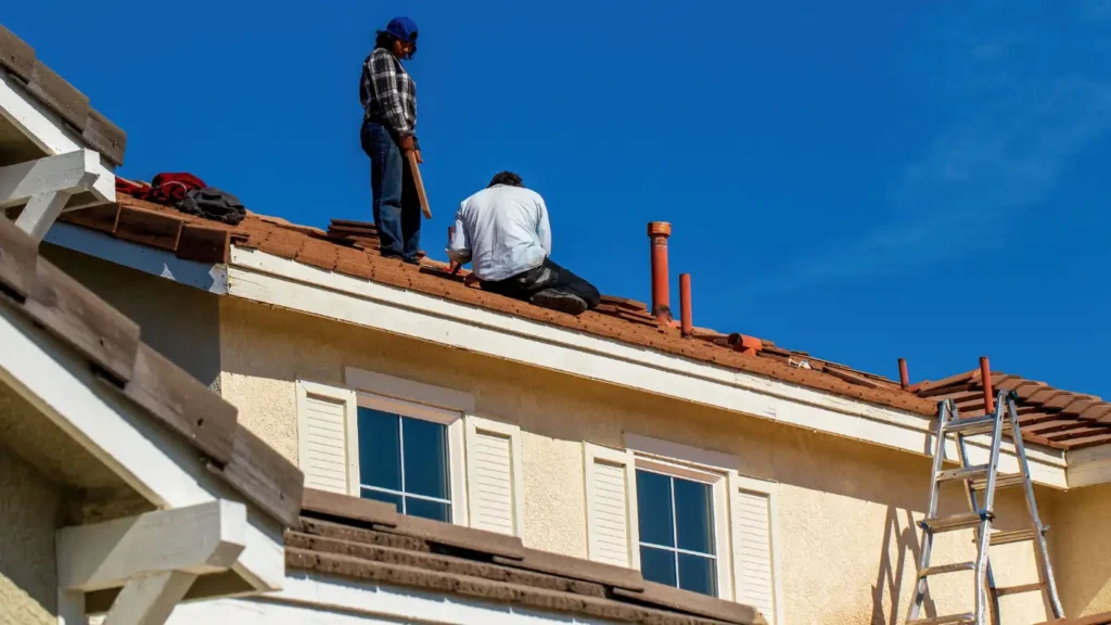 Two roofers working on a tile roof of a two-story stucco home under a clear blue sky, with an extension ladder against the gutter.