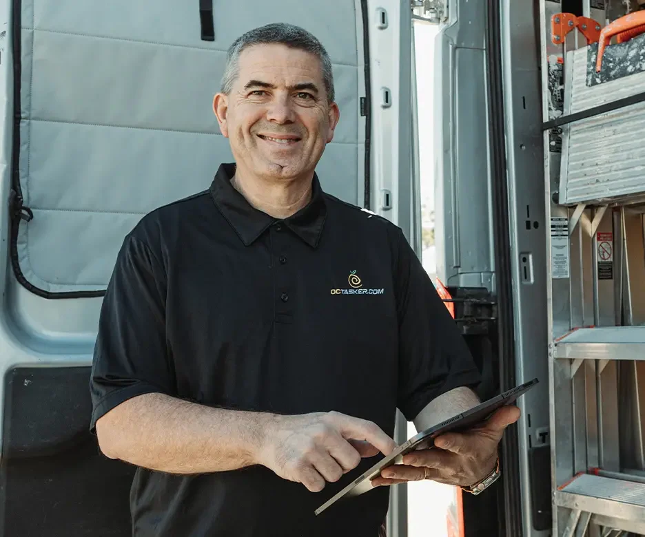 Smiling technician in a black polo with the OCTASKER.COM logo, standing beside a service van and holding a tablet.