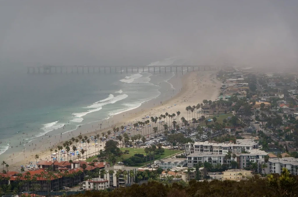 Aerial view of a foggy coastline with a long pier extending into the ocean, a sandy beach lined with palm trees, and buildings along the shore.