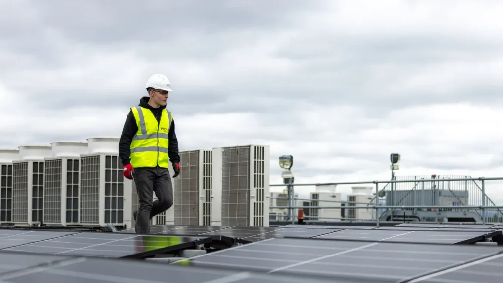 Worker in a high-visibility safety vest and hard hat walking on solar panels on a rooftop.