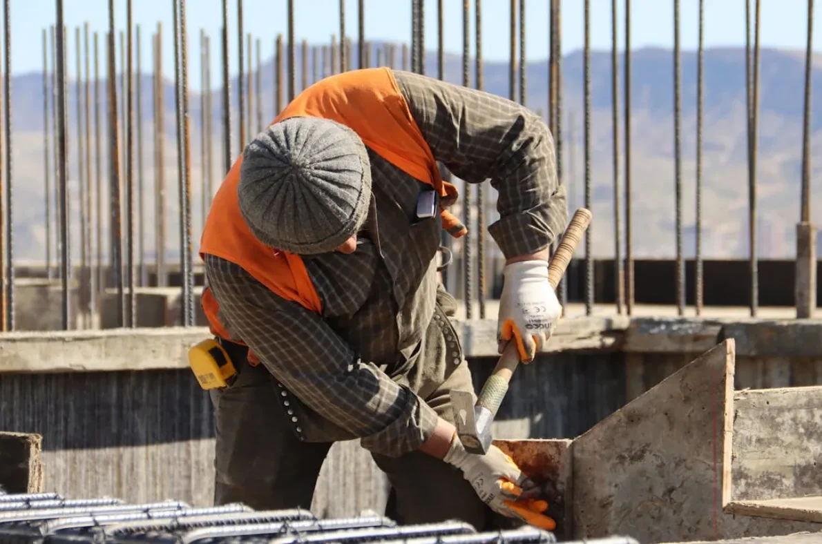Construction worker in a gray beanie and orange safety vest hammering near concrete formwork on a building site.