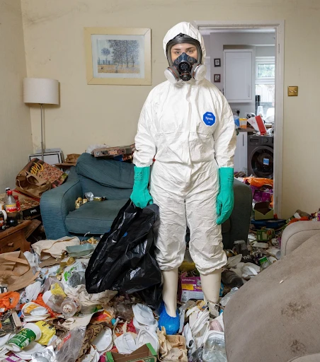 - Housecall Pro Person in a white hazmat suit with gas mask and green gloves stands in a cluttered living room, holding a black trash bag.
